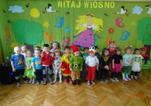Dzieci z grupy I w wiosennych przebraniach na tle dekoracji.